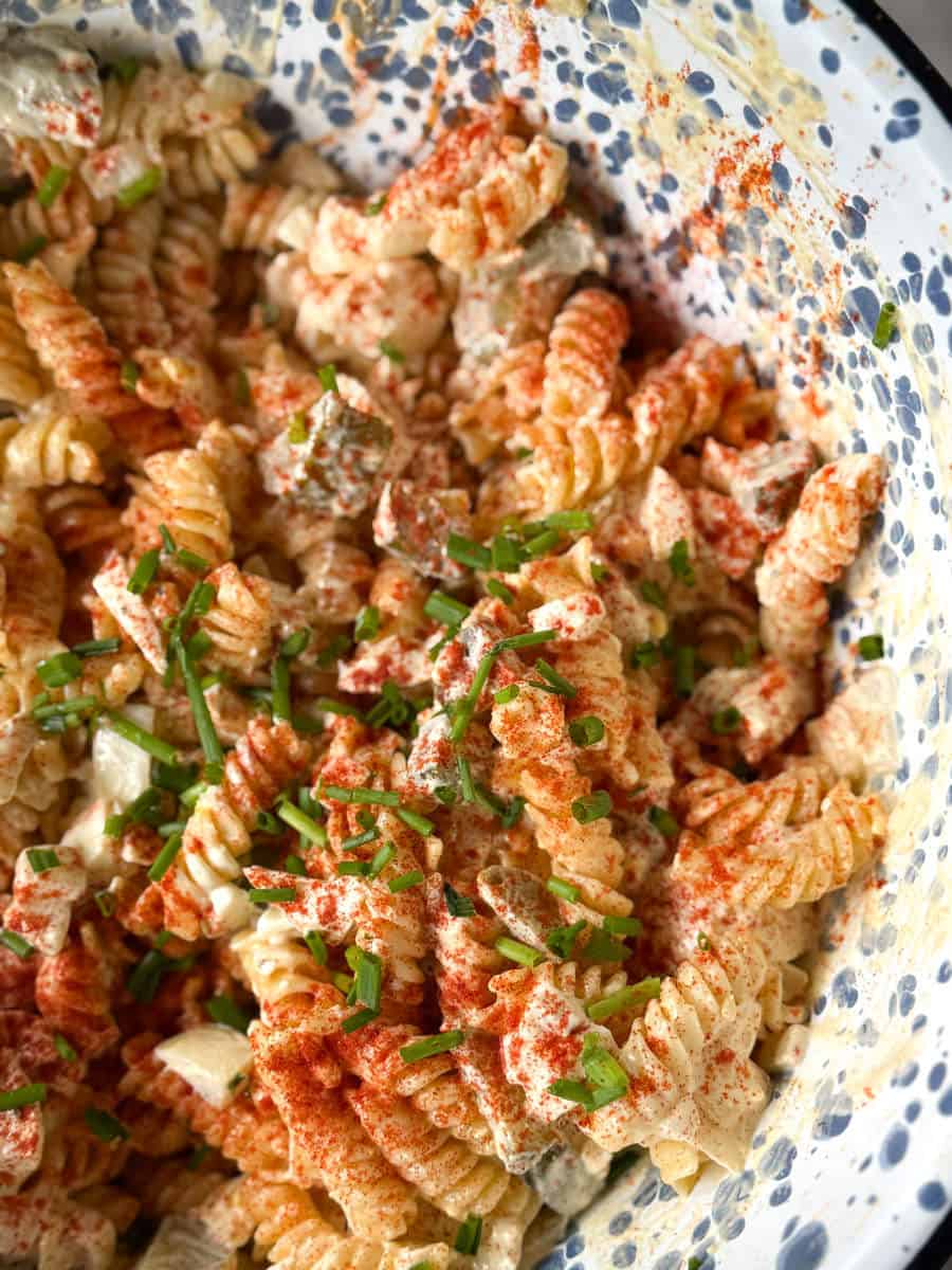 A close-up of a bowl of rotini pasta salad mixed with diced vegetables, creamy dressing, chopped chives, and sprinkled with paprika. The bowl has a speckled blue and white pattern.