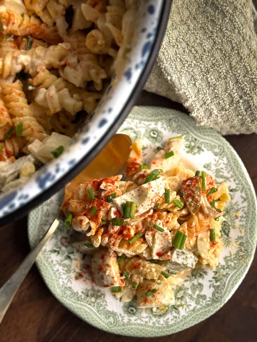 A plate of creamy pasta salad with diced vegetables and herbs, garnished with paprika and chopped chives, sits next to a serving dish with more salad. A spoon and textured cloth are also visible.