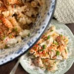 A close-up of a pasta salad with rotini noodles, diced vegetables, and a creamy dressing, topped with chopped chives and paprika, served in a bowl and on a decorative plate.