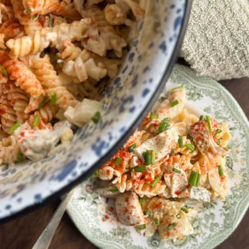 A close-up of a pasta salad with rotini noodles, diced vegetables, and a creamy dressing, topped with chopped chives and paprika, served in a bowl and on a decorative plate.