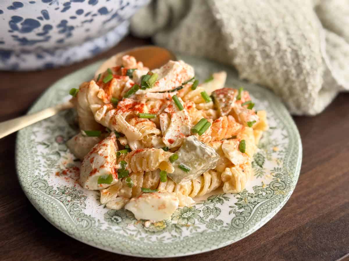A plate of creamy pasta salad with rotini, chopped vegetables, green onions, and a sprinkle of paprika, placed on a green floral-patterned dish next to a gold fork and a textured light green cloth.