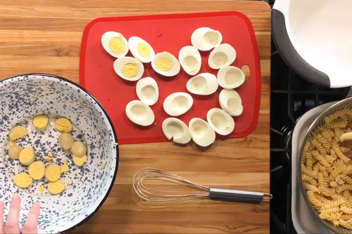 A cutting board with halved hard-boiled eggs, some with yolks removed. Egg yolks are in a bowl to the left. A whisk is on the counter, and a pot of cooked rotini pasta is on the right.