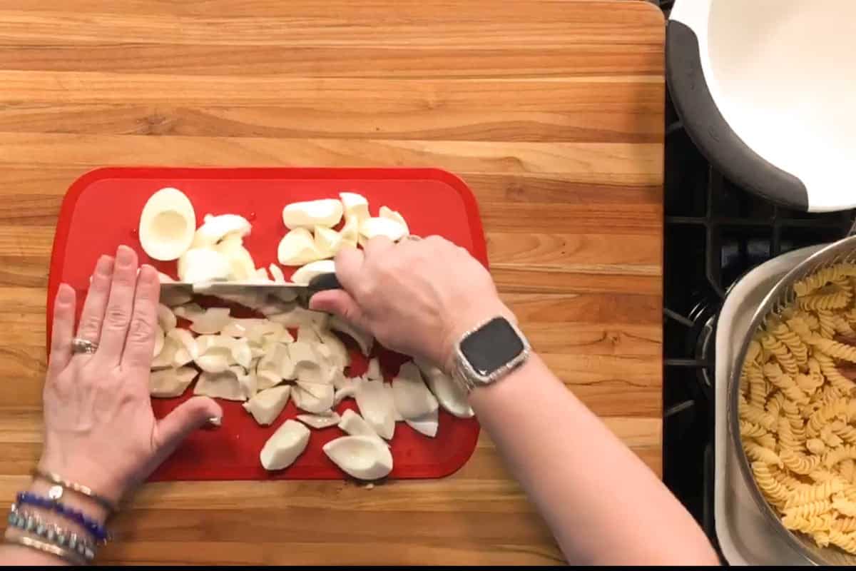 A person slices hard-boiled eggs on a red cutting board next to a stove with a pot of cooked rotini pasta. The person's left hand wears multiple bracelets and a smartwatch.