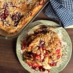 A plate with a serving of fruit crumble topped with streusel sits next to a glass baking dish with more crumble on a wooden surface; a spoon and a blue checkered cloth are nearby.