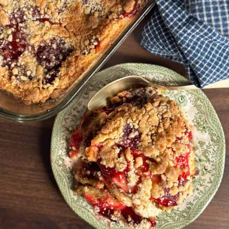A plate with a serving of fruit crumble topped with streusel sits next to a glass baking dish with more crumble on a wooden surface; a spoon and a blue checkered cloth are nearby.