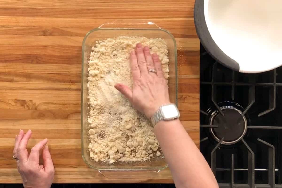 A person presses crumbly dough into a glass baking dish on a wooden countertop. Their left hand is visible, and they are wearing a watch on their right wrist. A stove and a white bowl are nearby.