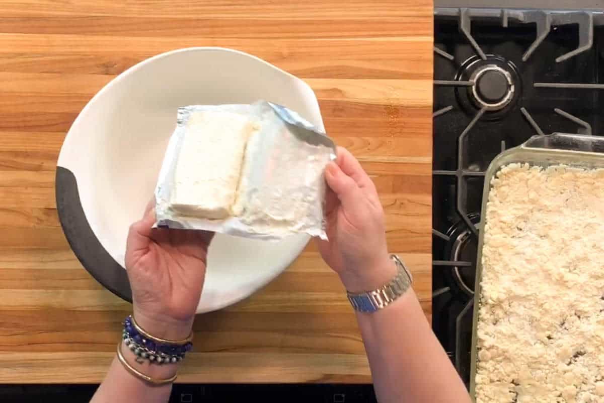 A person holds an opened package of cream cheese over a large empty white mixing bowl on a wooden countertop next to a stove and a glass baking dish filled with a crumbly mixture.