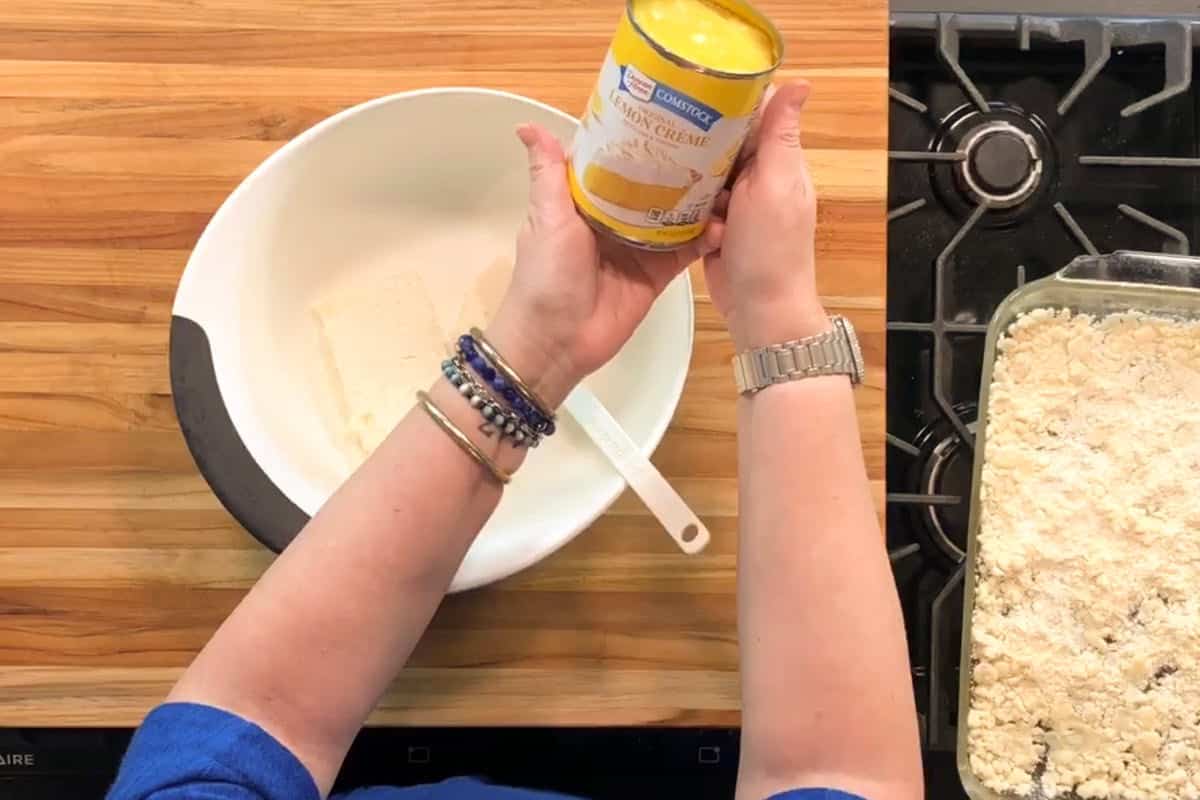 A person holds a can of sweetened condensed milk above a white mixing bowl containing cream cheese. A spatula rests in the bowl. A glass baking dish with a crumbly topping sits next to a gas stove on a wooden countertop.