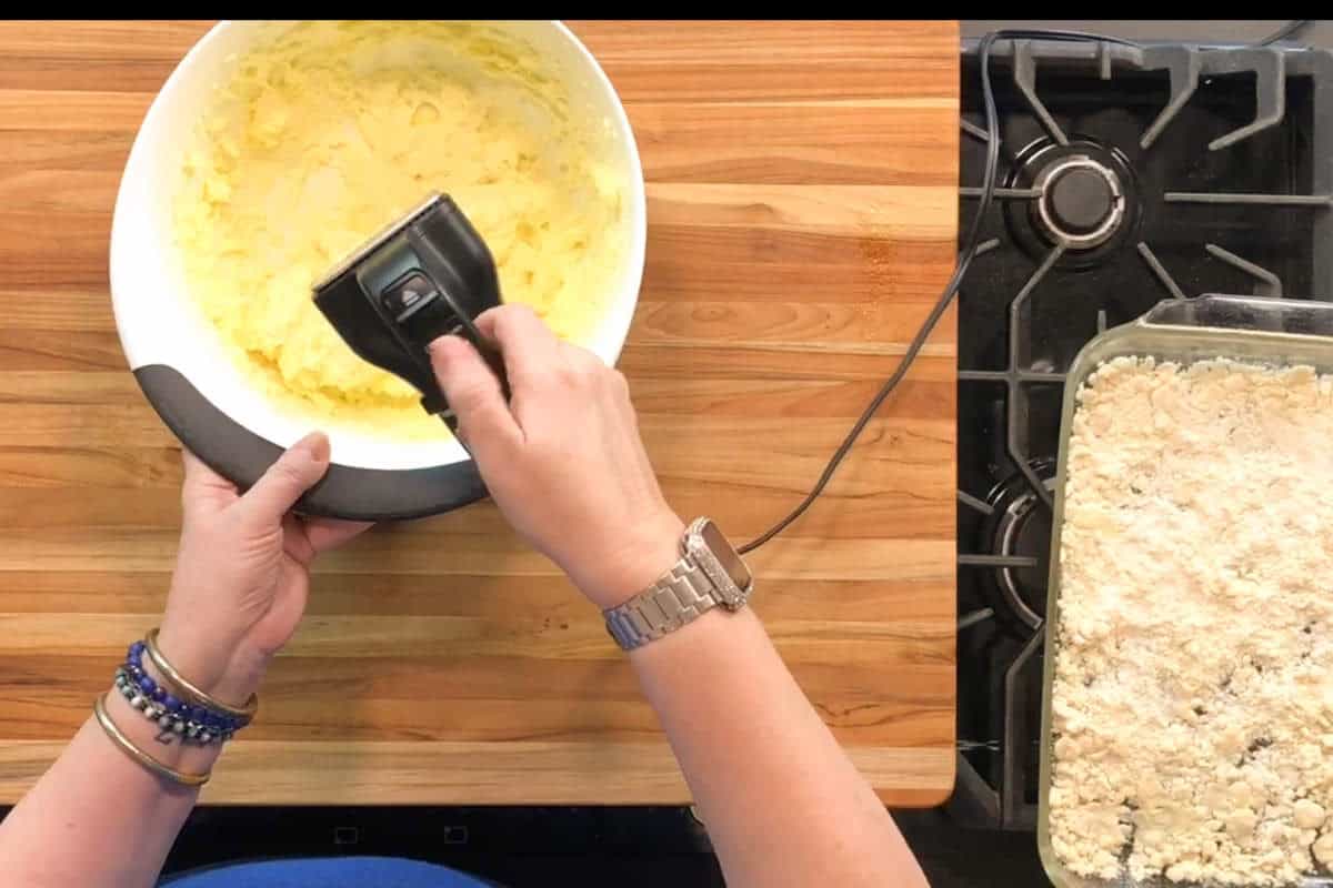 A person uses a hand mixer to blend a yellow batter in a white bowl on a wooden countertop; a glass baking dish with a crumbly mixture sits beside a stove.