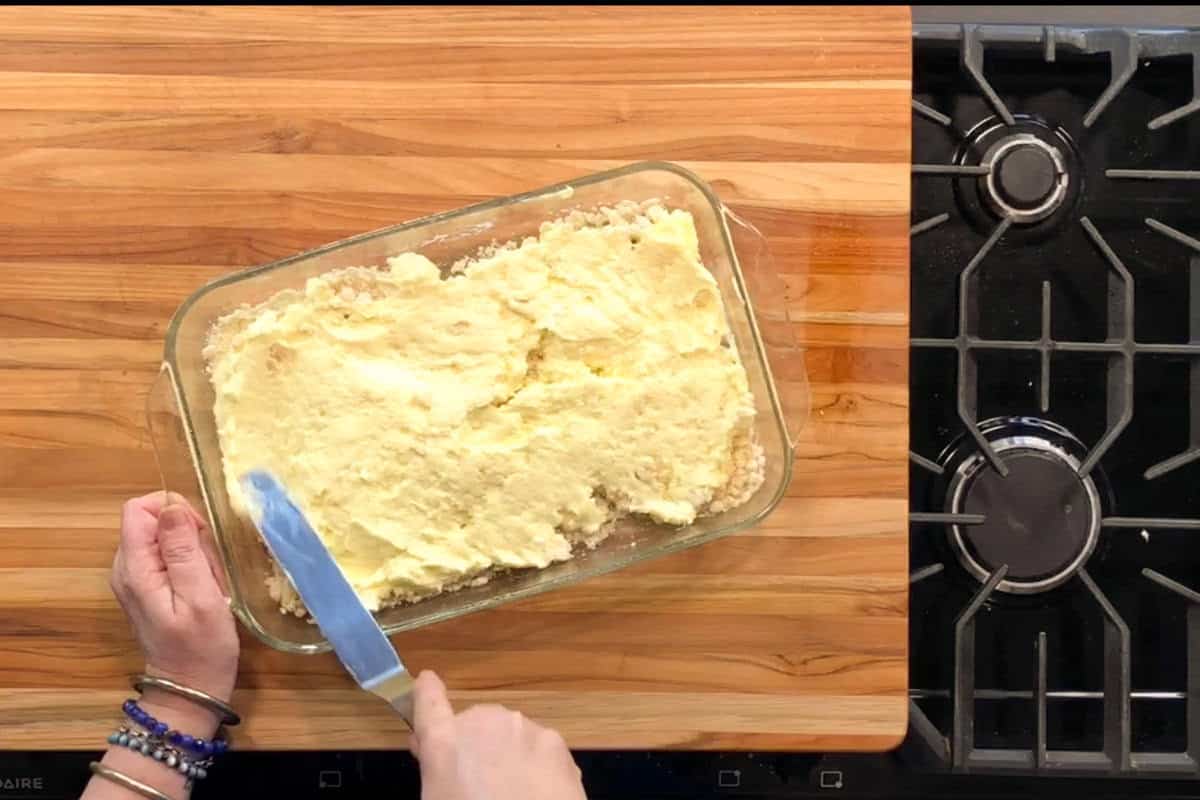 A person spreads a yellow batter or dough in a glass baking dish with a knife on a wooden countertop, next to a black gas stove.