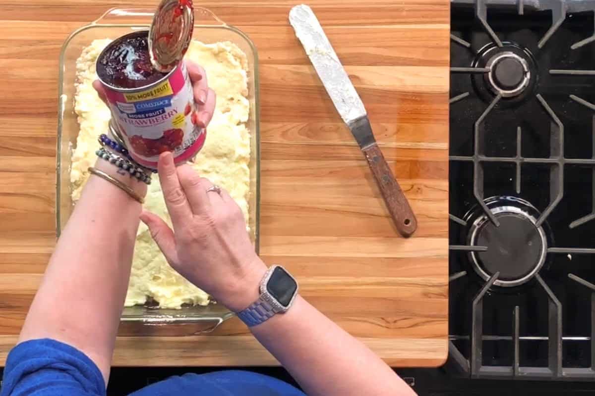 A person holding a can of strawberry filling above a baking dish with batter on a wooden counter; a metal spatula rests nearby next to a stovetop.