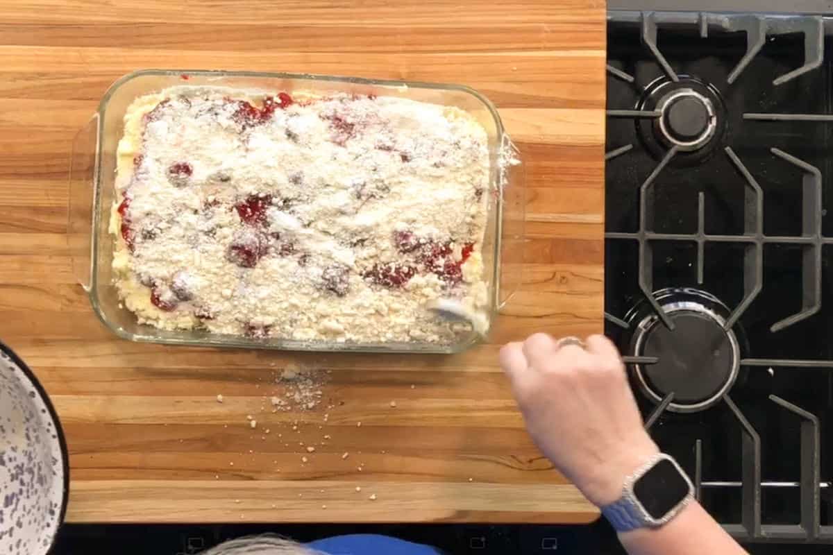 A person spreads crumb topping over a rectangular berry dessert in a glass baking dish on a wooden countertop next to a black gas stove. The person is wearing a blue shirt and a smartwatch.