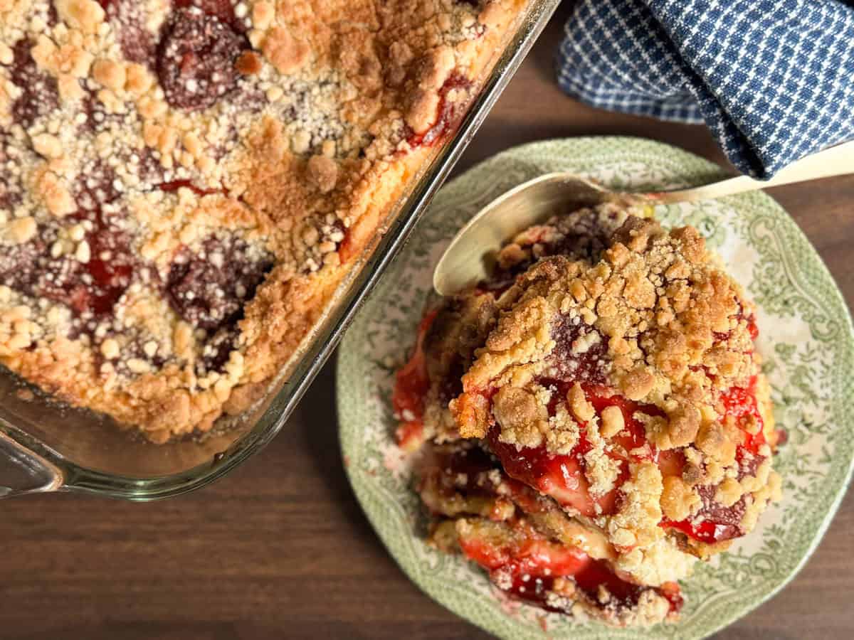 A serving of fruit crumble with a crumbly topping sits on a green patterned plate next to a baking dish containing more of the dessert. A metal spoon and blue checkered cloth are visible.