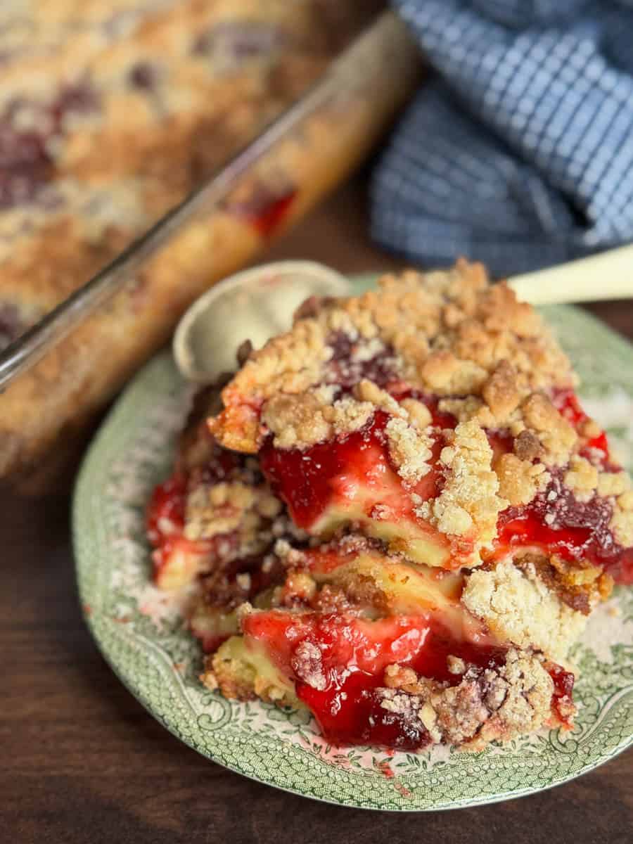 A close-up of a plate with a serving of cherry crumb dessert topped with crumbly streusel, with a spoon on the side and the remaining dessert and a blue cloth in the background.