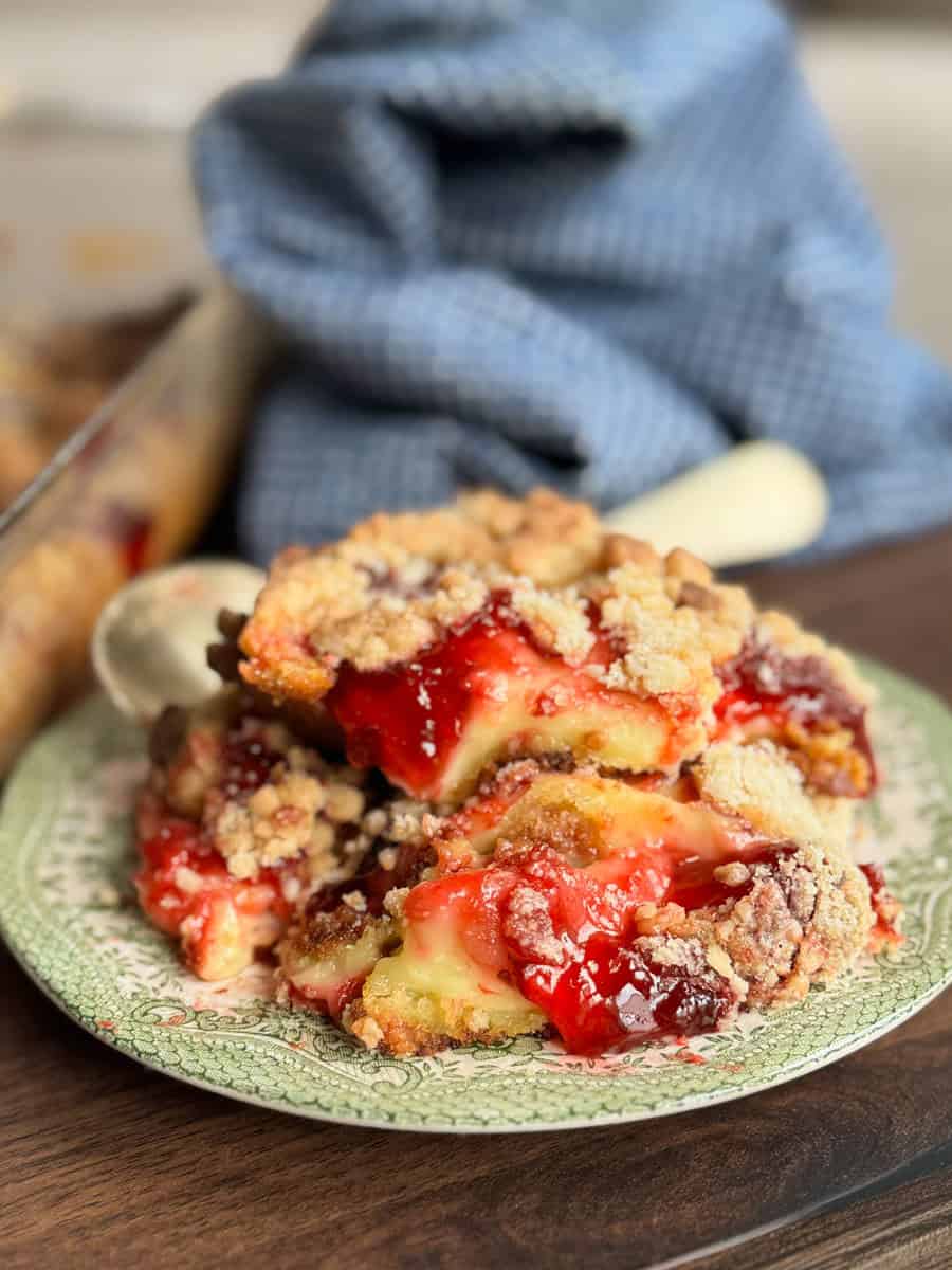Two slices of crumb-topped fruit bars with a red and yellow filling are stacked on a patterned plate, with a baking dish and blue towel in the background. The bars have a crumbly texture and are slightly oozing filling.