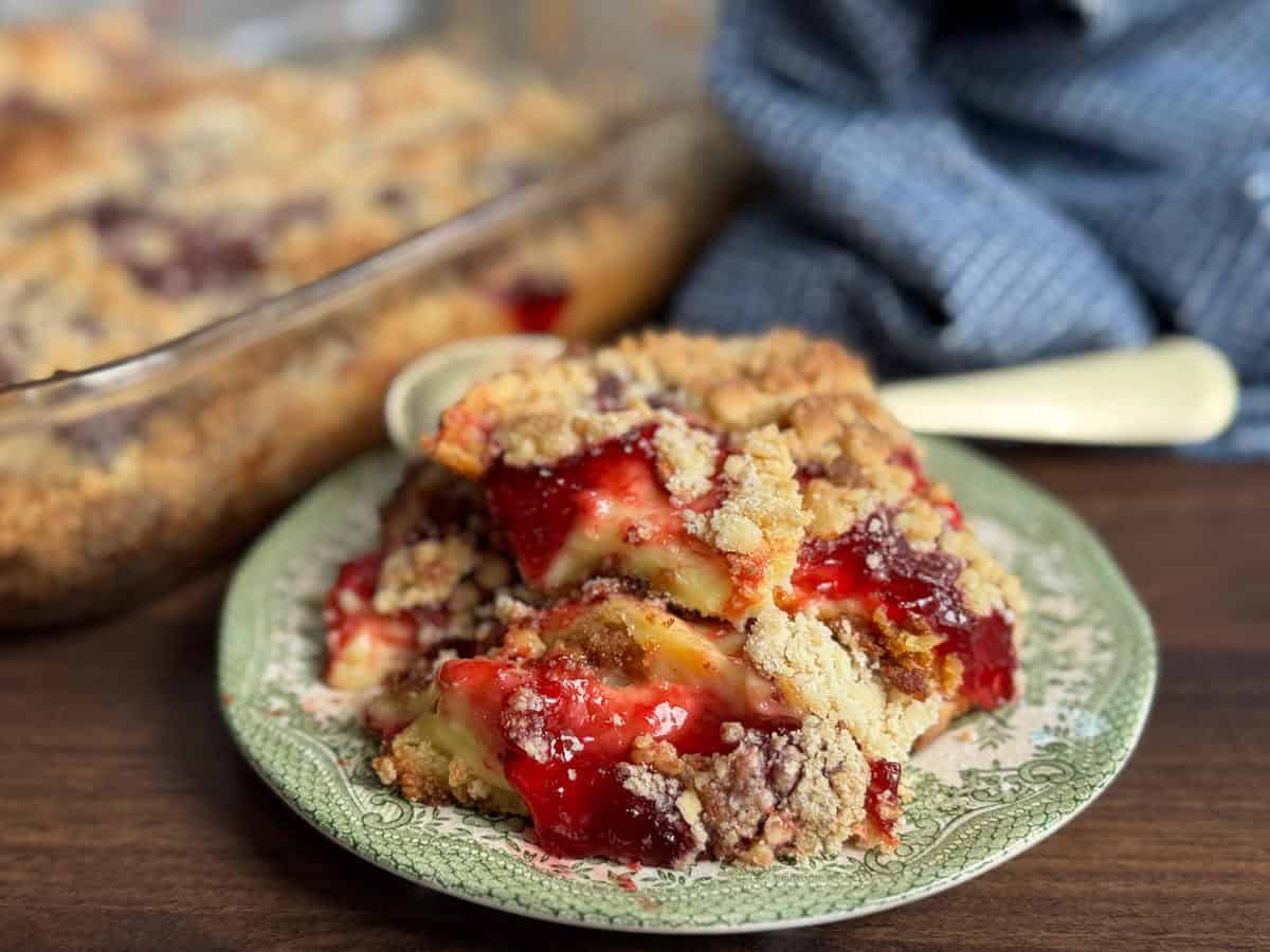 A serving of fruit crumble with a golden crumb topping and red fruit filling sits on a patterned plate with a spoon. More crumble is visible in a glass baking dish in the background. A blue cloth is nearby.