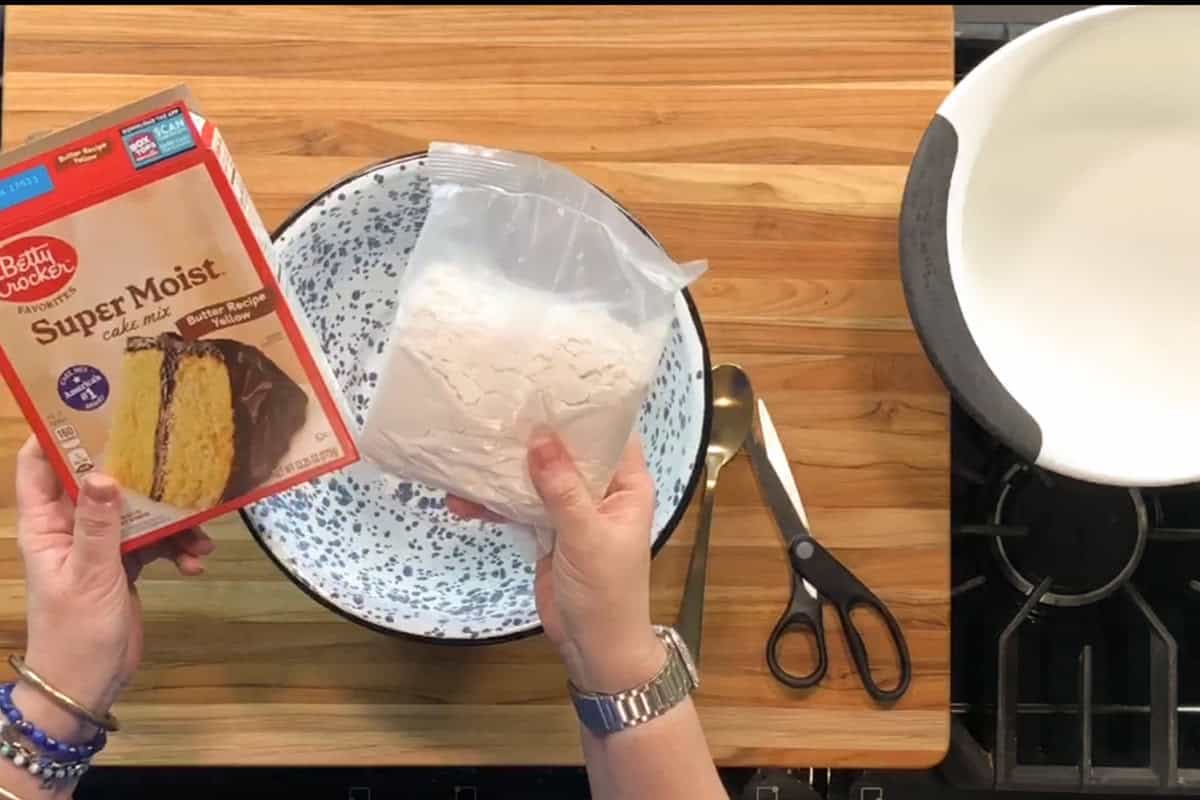 A person holds a box of Betty Crocker Super Moist yellow cake mix and an opened plastic bag of cake mix powder over a speckled mixing bowl on a wooden countertop, with scissors and a spoon beside the bowl.