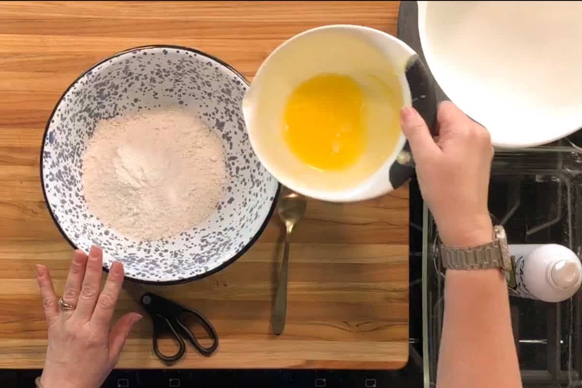 A person holds a bowl of melted butter above a large bowl containing flour mixture on a wooden countertop. A pair of scissors, a spoon, and a can of cooking spray are nearby.