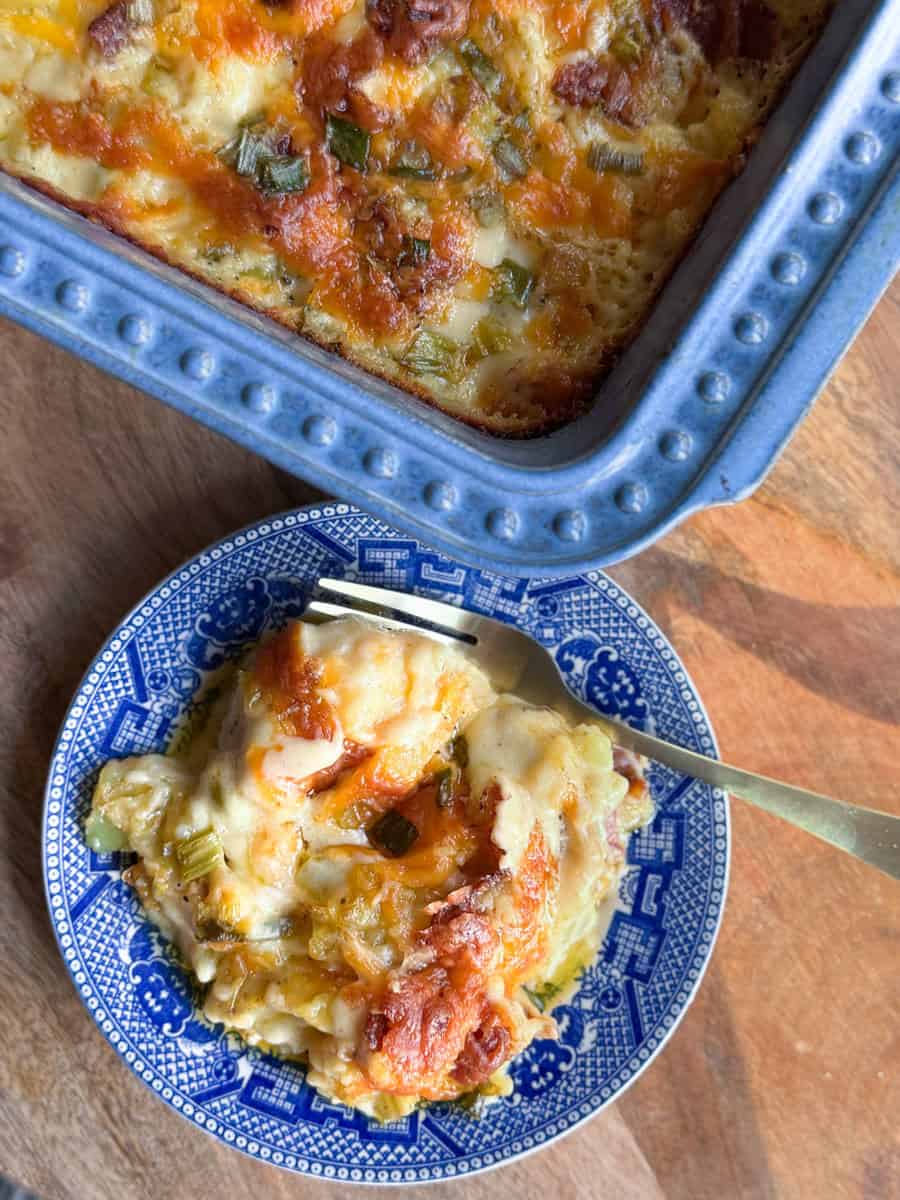 A serving of cheesy casserole with green onions on a blue patterned plate next to a blue baking dish containing the rest of the casserole, placed on a wooden surface. A fork rests on the plate.