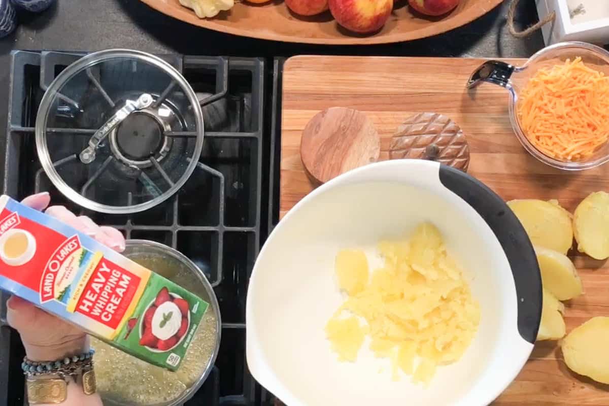 A person pours heavy whipping cream from a carton into a large white mixing bowl containing cooked potatoes. Sliced potatoes, shredded cheese, and a covered pot are on the surrounding counters.
