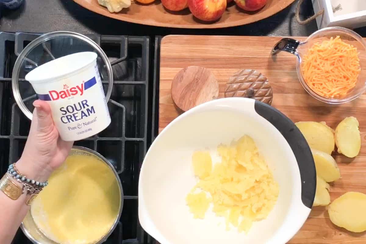 A hand holds a container of Daisy sour cream over a kitchen counter with a bowl of mashed potatoes, a bowl of beaten eggs, shredded cheddar cheese, and sliced potatoes on a cutting board. Apples are visible in the background.