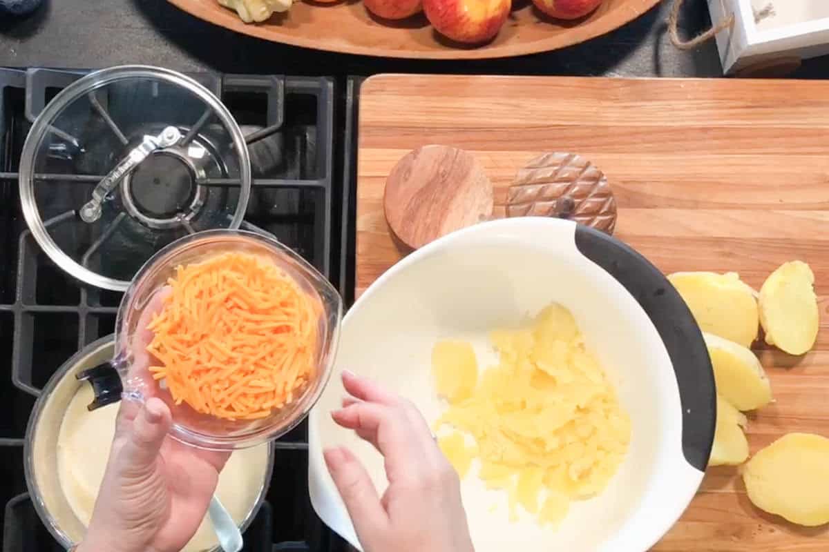 A person holds a bowl of shredded orange cheese over a white mixing bowl with sliced potatoes. Nearby, a pot sits on the stove and more potato slices are on a wooden cutting board.
