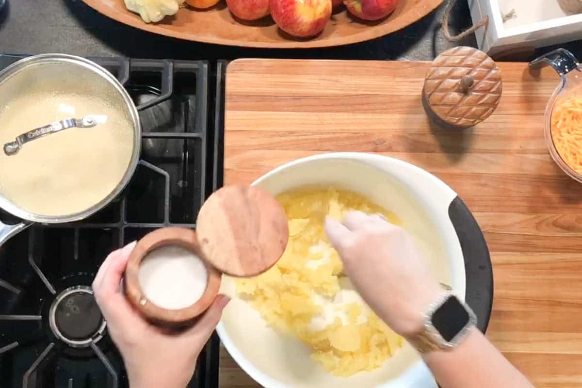 A person adds sugar from a wooden container into a white mixing bowl with yellow batter on a wooden cutting board, next to a saucepan on the stove and a bowl of shredded carrots.