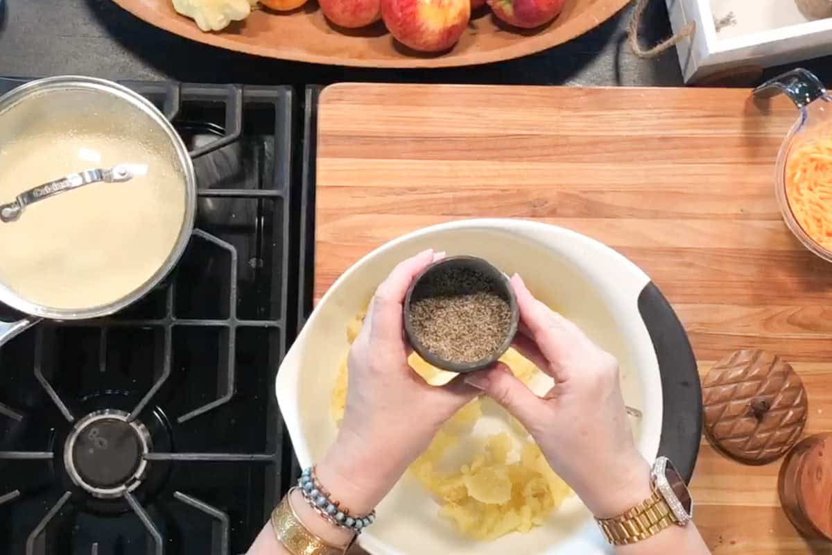 Overhead view of hands holding a small bowl of ground pepper above a mixing bowl with mashed potatoes, next to a stove with a pot and a wooden cutting board with apples and grated cheese.