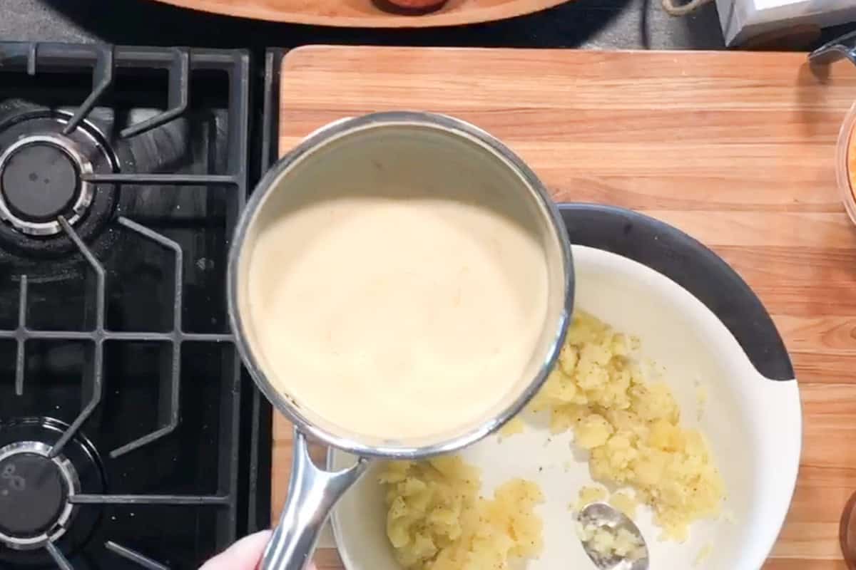 A hand holds a saucepan with a creamy liquid above a bowl containing mashed potatoes. The scene is set on a wooden cutting board beside a gas stove. A spoon rests in the bowl.