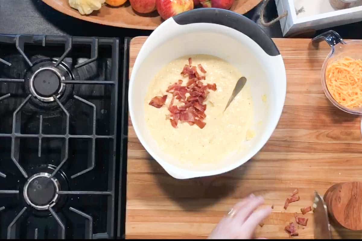 A mixing bowl filled with thick batter and bacon pieces sits on a wooden cutting board. A spoon is in the bowl, shredded cheese is nearby, and a cooktop is visible on the left side of the image.
