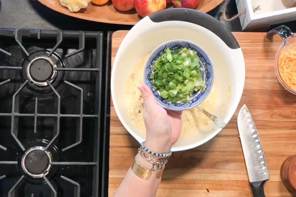 A person holds a bowl of chopped green onions above a mixing bowl of batter on a wooden countertop next to a kitchen knife, grated cheese, and a gas stove.