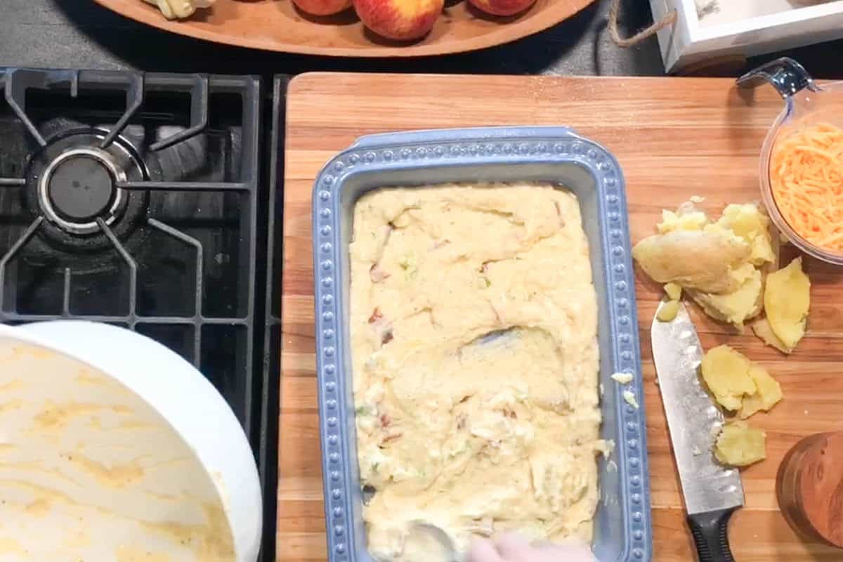 A baking pan with uncooked batter is on a wooden cutting board next to a gas stove. Nearby are a knife, peeled and unpeeled potatoes, grated cheese in a cup, and a bowl with leftover batter.