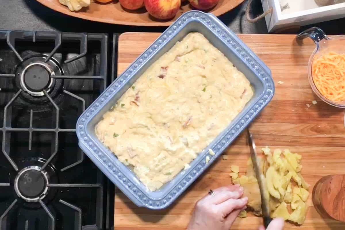A person chops cooked potatoes on a wooden cutting board next to a blue baking dish filled with batter, a cup of shredded cheese, a stove, and a tray of peaches.