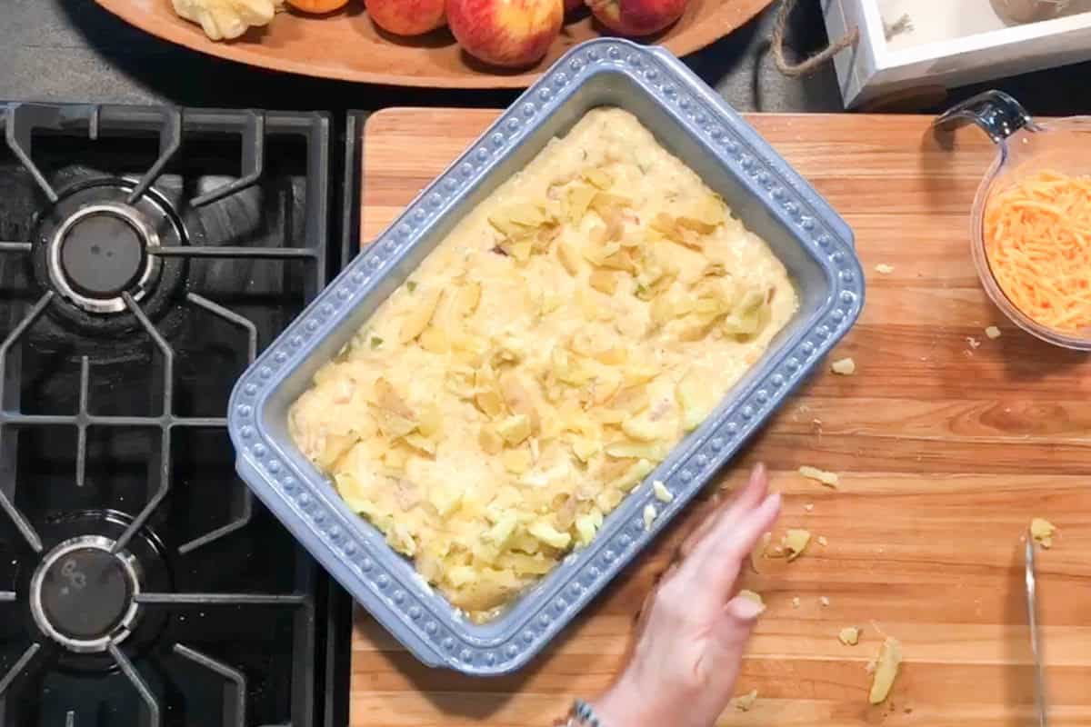 A rectangular blue baking dish filled with an unbaked casserole topped with crushed potato chips sits on a wooden cutting board near a stove, with a hand visible in the lower right corner.