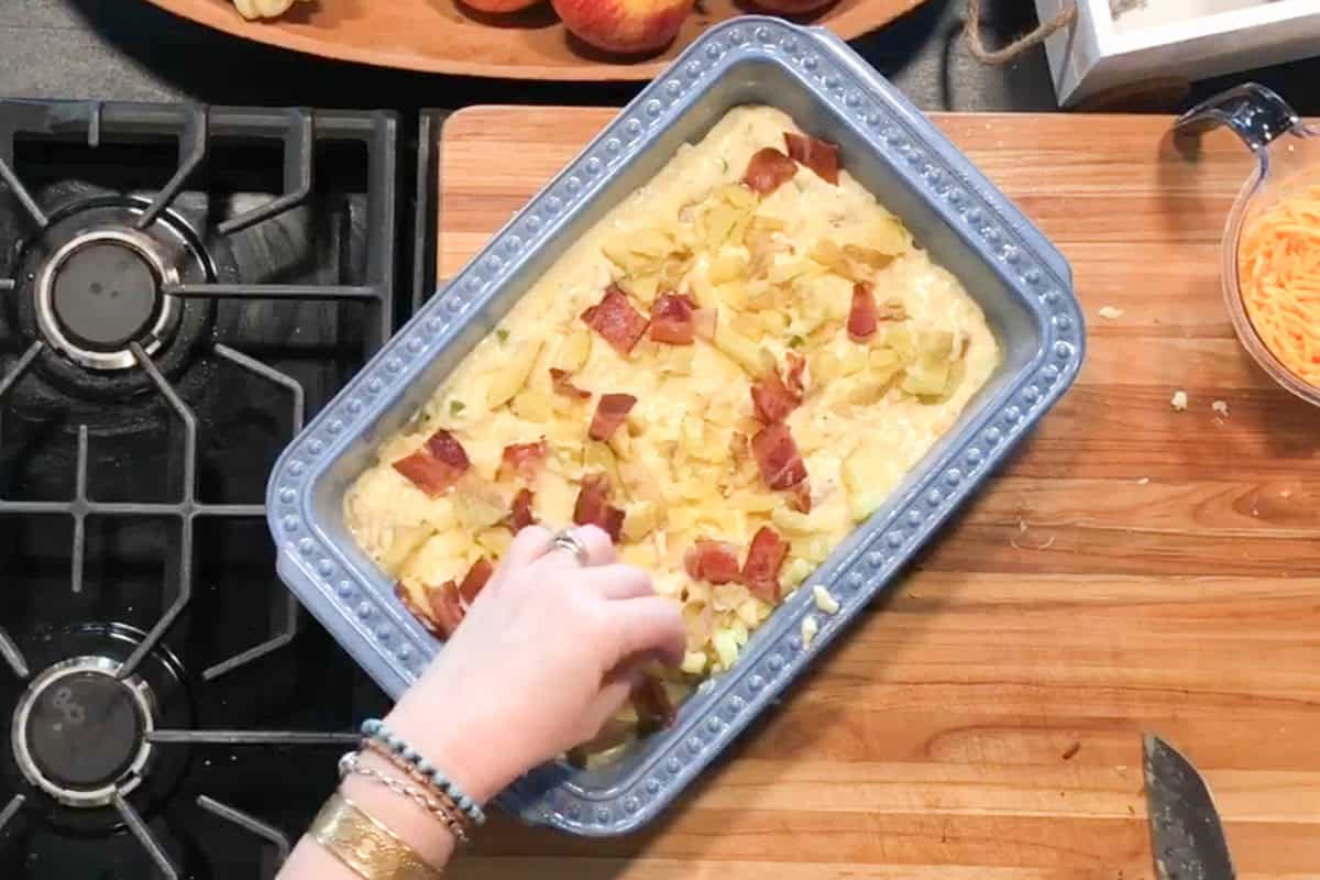 A person’s hand sprinkles chopped ingredients, possibly bacon and potatoes, onto a casserole in a blue baking dish on a wooden counter next to a gas stove and a bowl of shredded cheese.