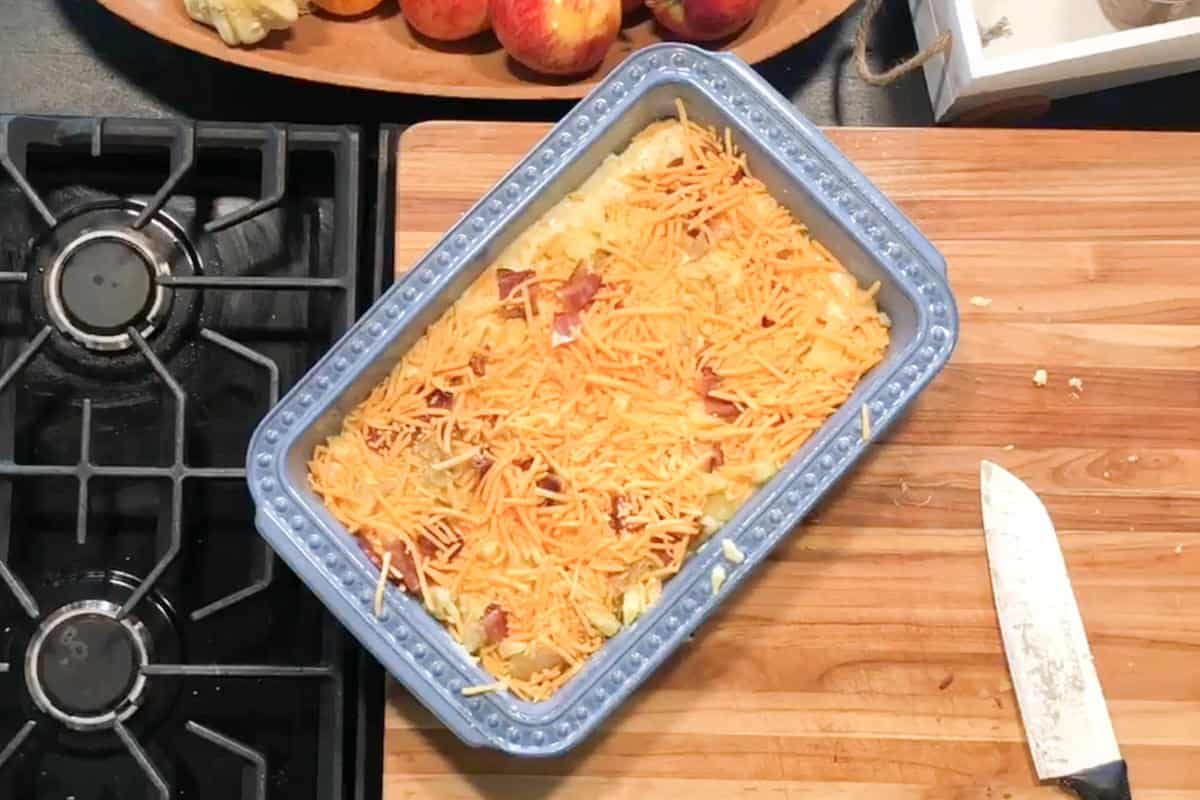 A blue baking dish filled with an unbaked casserole topped with shredded cheese sits on a wooden cutting board next to a kitchen knife. A stovetop and a bowl of apples are partially visible nearby.