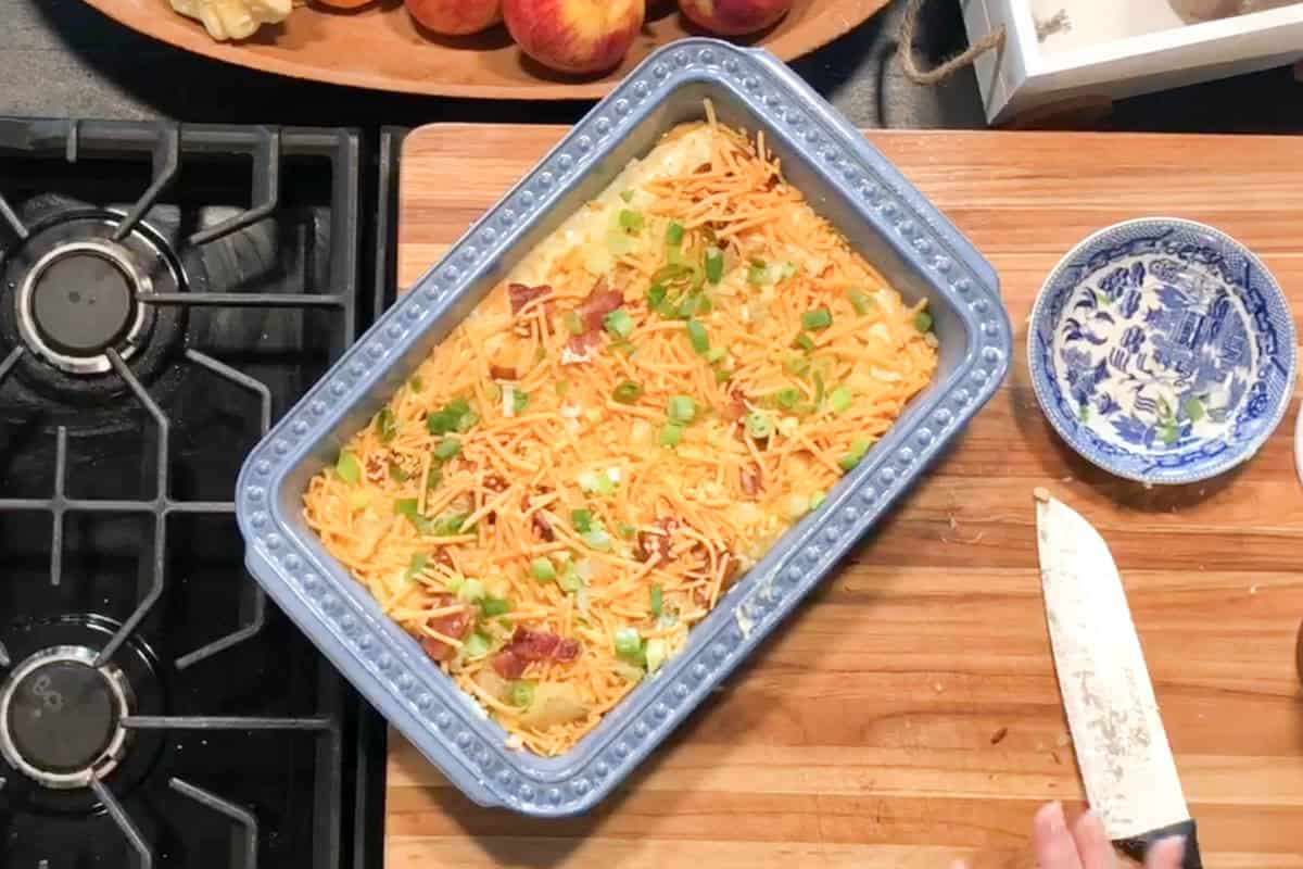 A blue baking dish filled with a casserole topped with shredded cheese and chopped green onions sits on a wooden cutting board next to a stove. A hand holds a knife beside a small blue-and-white bowl.