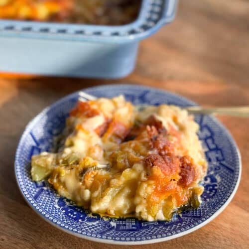 A serving of baked casserole with a golden, melted cheese topping is on a blue and white patterned plate. Part of the casserole dish is visible in the background.