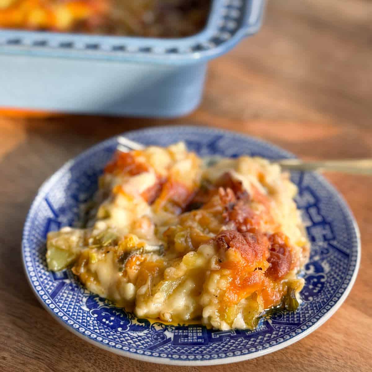 A serving of baked casserole with a golden, melted cheese topping is on a blue and white patterned plate. Part of the casserole dish is visible in the background.