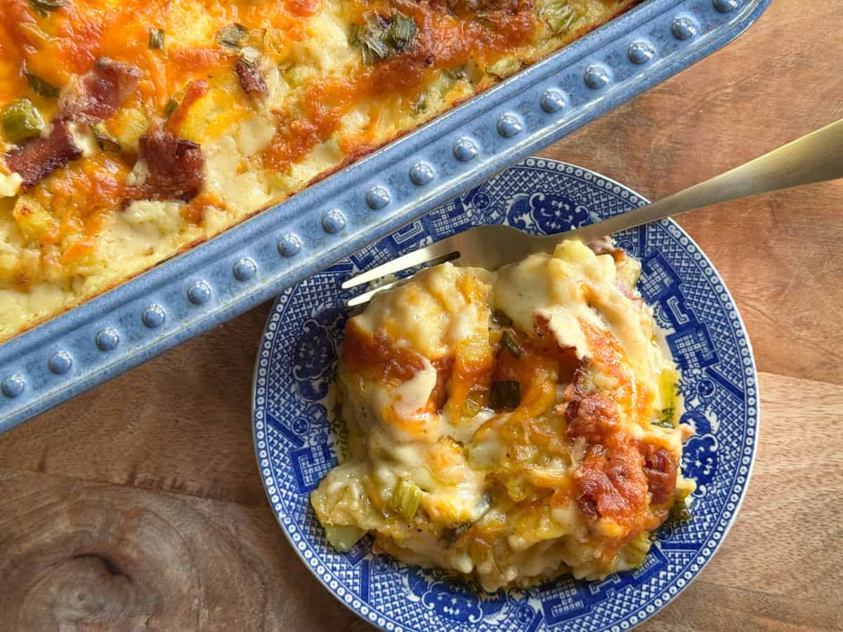 A portion of cheesy casserole with browned top is served on a blue patterned plate, next to a baking dish containing the rest of the casserole, on a wooden surface. A fork rests on the plate.