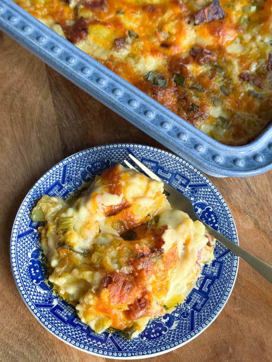 A serving of cheesy casserole with visible chunks of bread and vegetables on a blue patterned plate, with a fork. The casserole dish with the remaining portion is partially visible on a wooden surface.