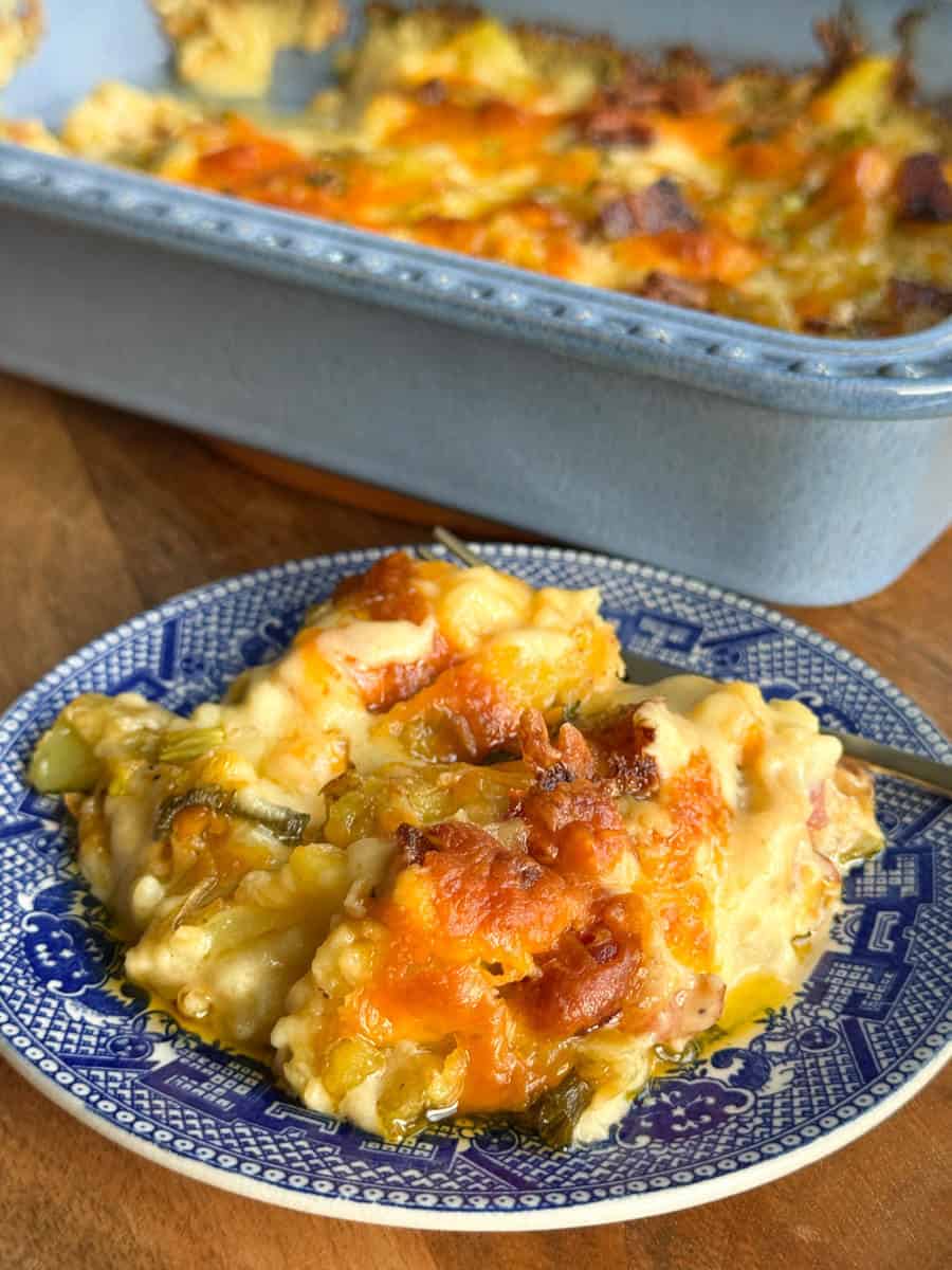 A serving of cheesy casserole with a browned top sits on a blue and white patterned plate, with a baking dish containing more casserole in the background.