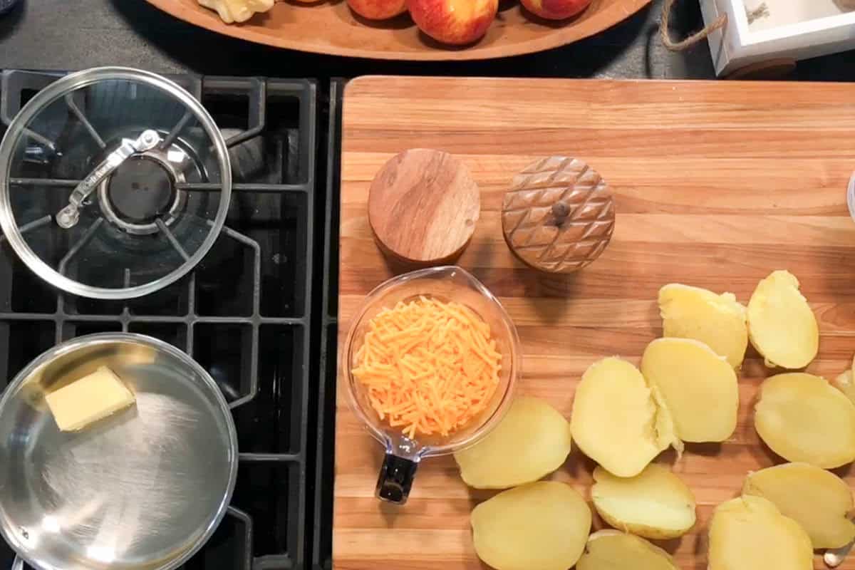 A stove with a pan of butter, a cutting board with halved boiled potatoes, a measuring cup of shredded cheddar cheese, wooden salt and pepper shakers, and a tray of apples in the background.