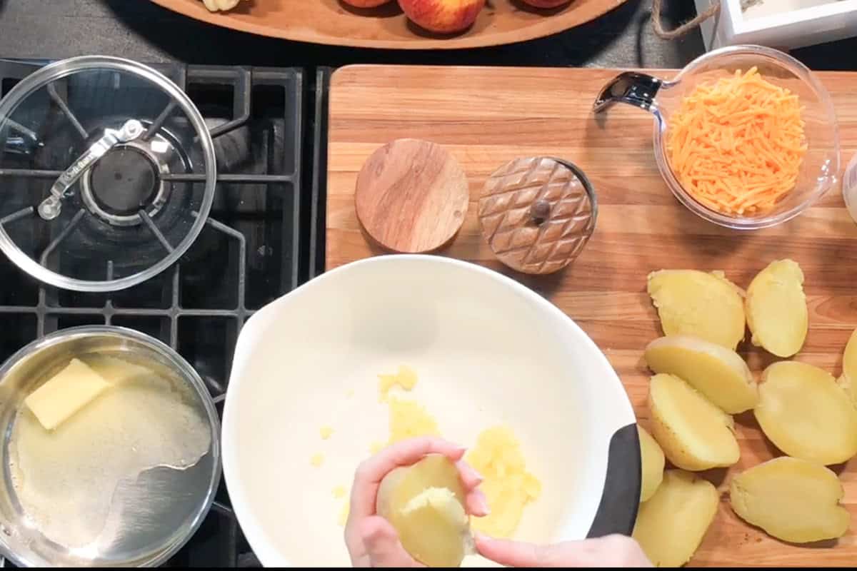 A person scoops the insides from boiled potato halves into a white mixing bowl. A saucepan with melted butter, shredded cheese, salt, and pepper are arranged on a wooden cutting board nearby.