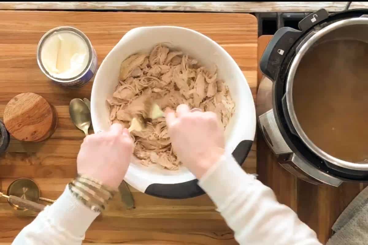 A person mixes shredded chicken in a white bowl on a wooden countertop. An open can of cream, two spoons, seasonings, and an Instant Pot with broth are nearby.