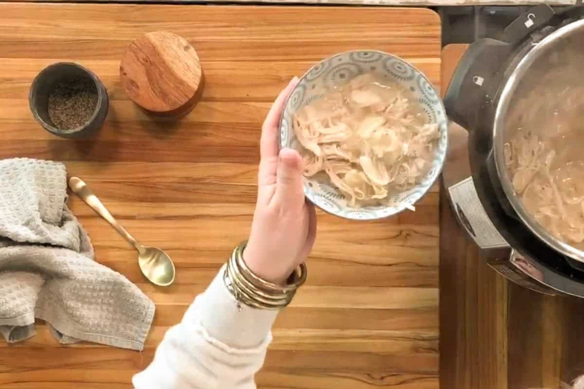 A hand holding a bowl of shredded chicken and broth above a wooden countertop with spices, a spoon, and a towel nearby; an electric pressure cooker sits to the right.