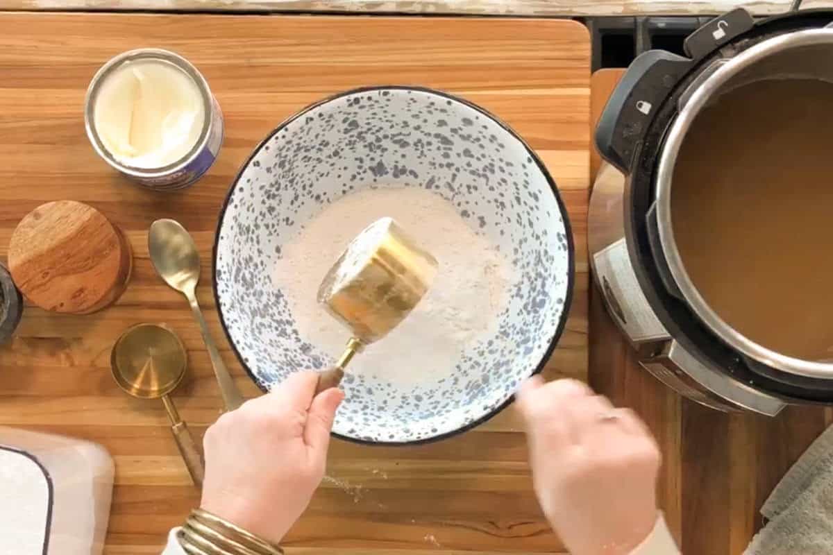 A person holds a gold measuring cup above a speckled mixing bowl with flour on a wooden countertop. Nearby are a can of shortening, two spoons, more measuring cups, and an Instant Pot with liquid inside.