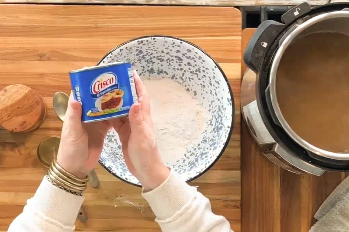 A person holds a can of Crisco shortening over a mixing bowl filled with flour on a wooden countertop. Measuring spoons, a small container, and an electric pressure cooker are nearby.