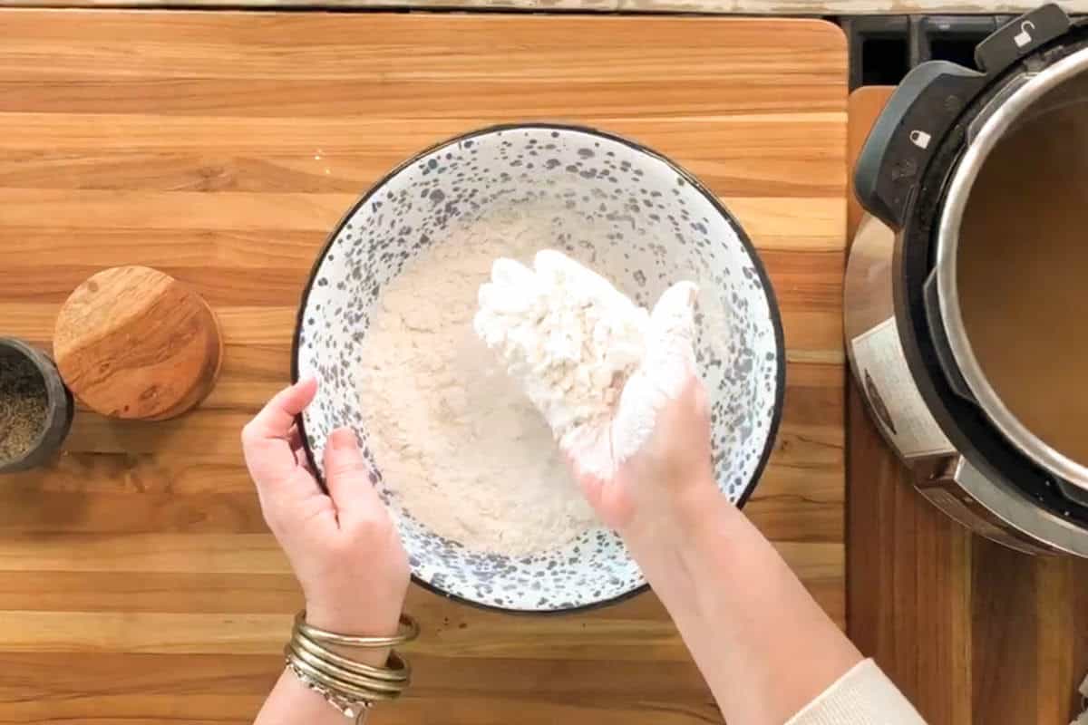A person holds a handful of flour mixture over a speckled bowl on a wooden countertop, wearing a white glove and several bracelets. A pressure cooker and a container of pepper are nearby.