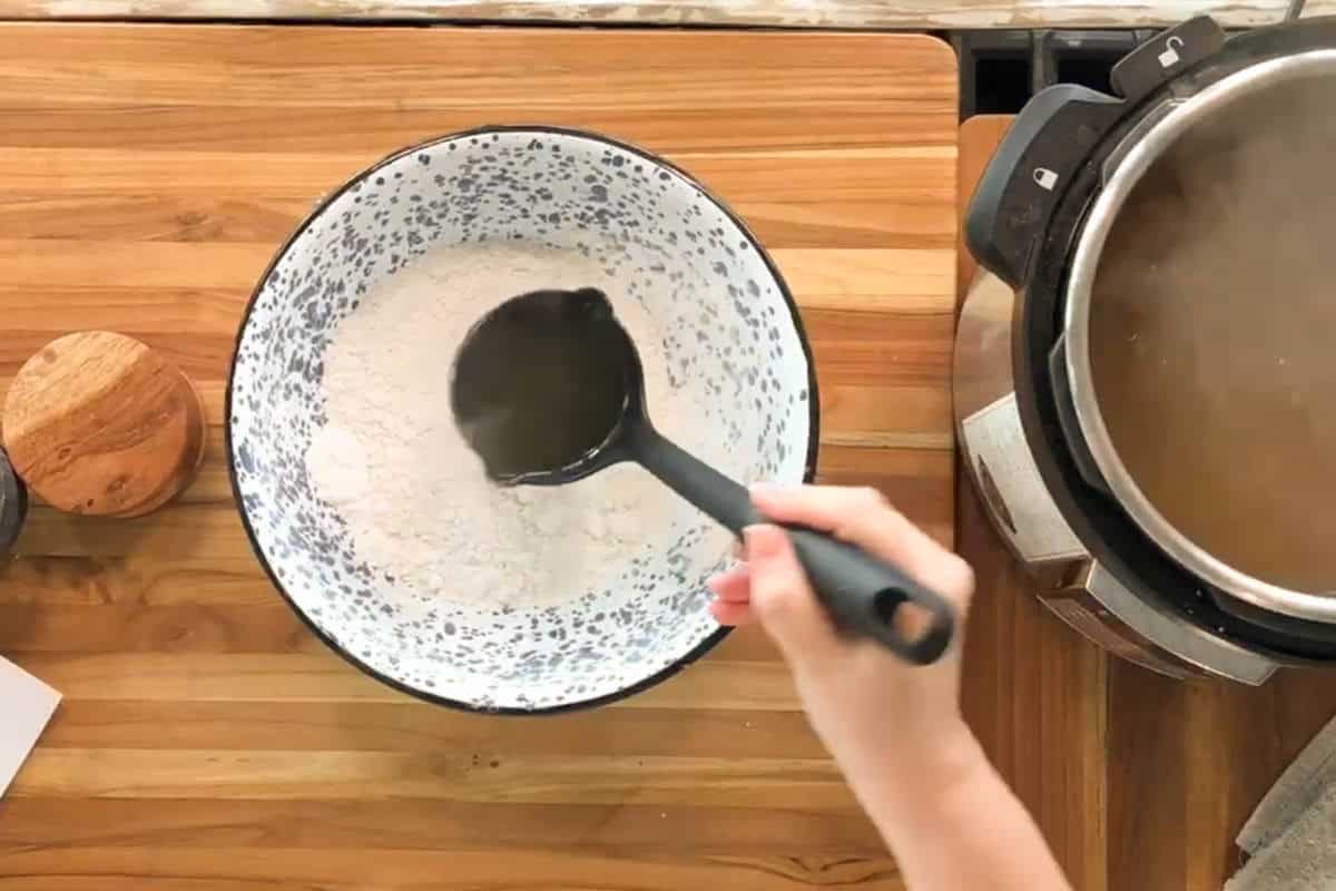 A hand is pouring hot liquid from a black ladle into a bowl filled with white flour mixture on a wooden countertop next to an electric pressure cooker.